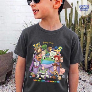 May include: A charcoal grey t-shirt featuring a colourful graphic of Toy Story characters emerging from a purple backpack. The design includes Buzz Lightyear, Woody, Rex, Mr. Potato Head, and others, with the text Toy Story 5 and You've Got A Friend In Me.
