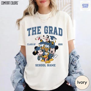 Puede incluir: Camiseta color marfil con la frase azul "THE GRAD", "CLASS OF 2026" y "SCHOOL NAME". Presenta un dise&ntilde;o de personajes de Disney con atuendo de graduaci&oacute;n. La camiseta es de Comfort Colors.