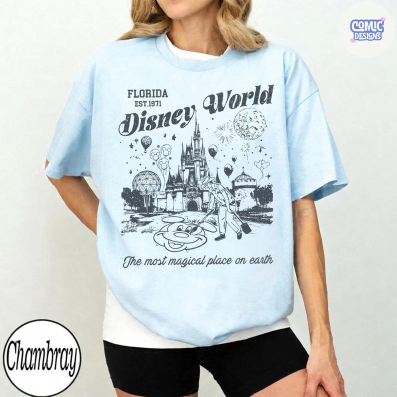K&ouml;nnte beinhalten: Hellblaues T-Shirt mit einem Disney World-Motiv, das Cinderella's Schloss und Feuerwerk zeigt. Der Text lautet "Florida Est.1971 Disney World" und "The most magical place on earth."
