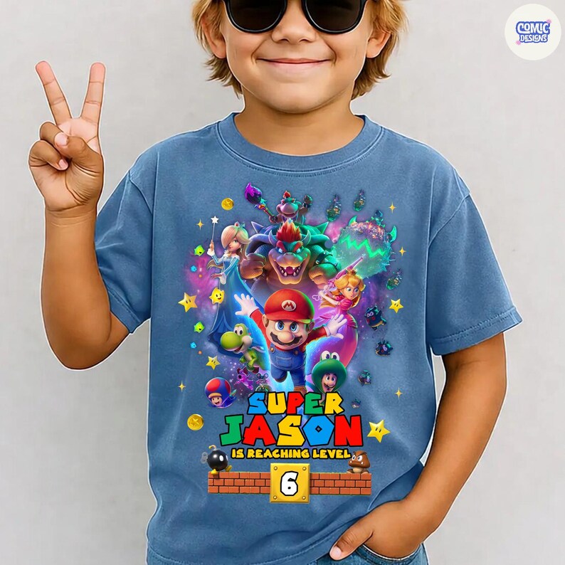 Pode incluir: Camiseta azul com um gr&aacute;fico do Super Mario com Mario e outros personagens. O texto "SUPER JASON IS REACHING LEVEL 6" est&aacute; impresso abaixo do gr&aacute;fico. A camiseta &eacute; usada por uma crian&ccedil;a.