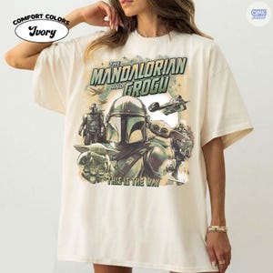 May include: Ivory-colored t-shirt featuring a graphic of The Mandalorian and Grogu. The design includes the text "The Mandalorian and Grogu" and "This is the Way", along with images of characters and spacecraft.