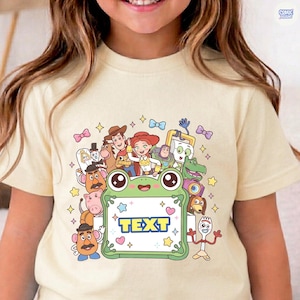 May include: A cream-colored t-shirt featuring a graphic print of various animated characters surrounding a central green frog-shaped frame. The frame contains the word TEXT in blue letters, accented by small stars and hearts on a white background.