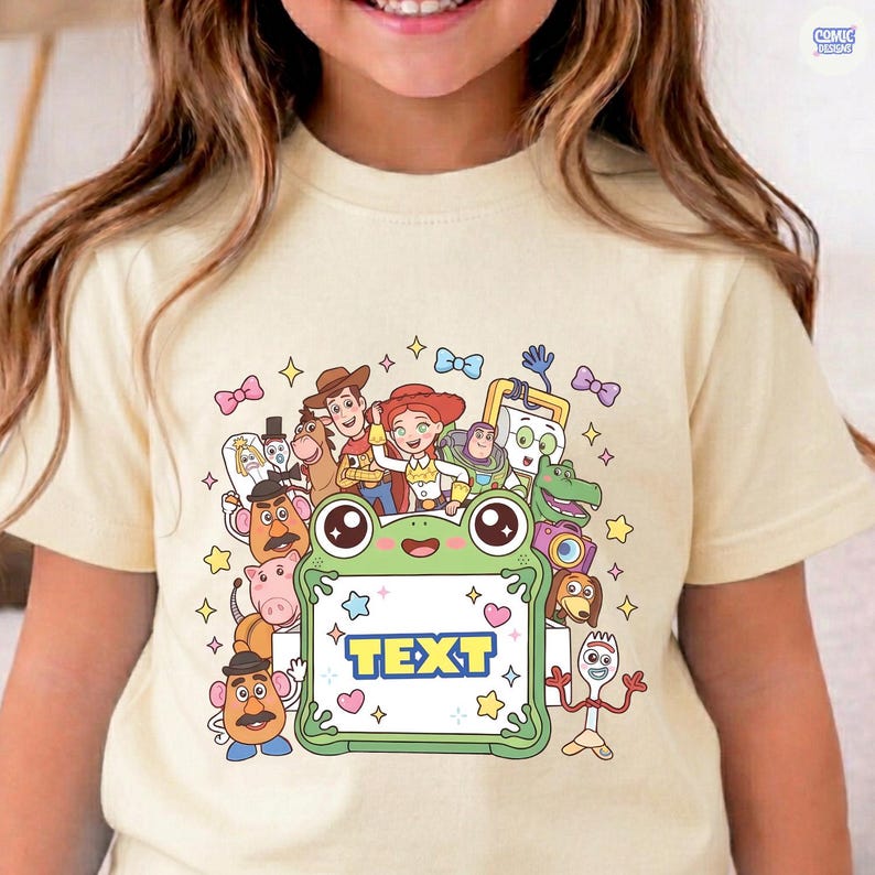 May include: A cream-colored t-shirt featuring a graphic print of various animated characters surrounding a central green frog-shaped frame. The frame contains the word TEXT in blue letters, accented by small stars and hearts on a white background.