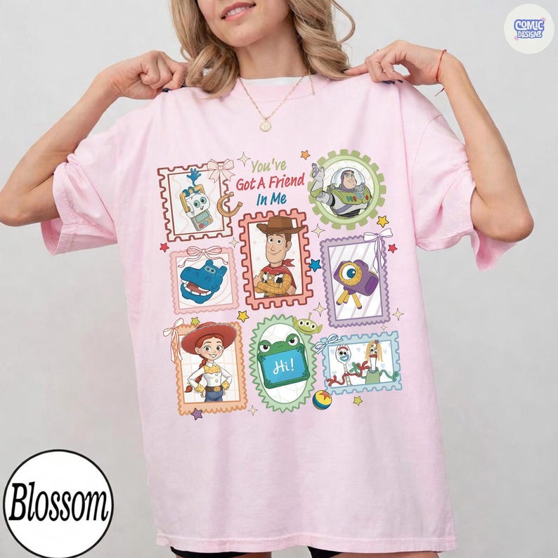May include: A light pink t-shirt featuring a graphic print of various Toy Story characters inside postage stamp-style frames. The text "You've Got A Friend In Me" is printed in the center. Characters include Woody, Buzz Lightyear, Jessie, and Forky.