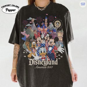 Puede incluir: Una camiseta gris carb&oacute;n de manga corta con un estampado gr&aacute;fico de personajes de dibujos animados con atuendo patri&oacute;tico frente a un castillo y fuegos artificiales. El texto dice Disneyland America 250. Color Pepper de Comfort Colors.