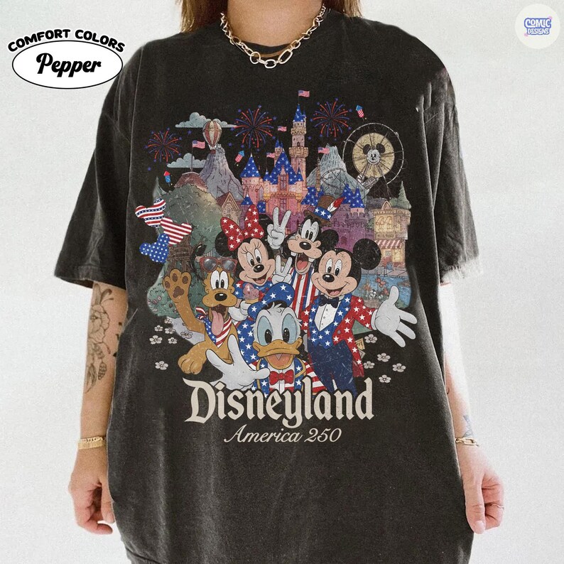 Puede incluir: Una camiseta gris carb&oacute;n de manga corta con un estampado gr&aacute;fico de personajes de dibujos animados con atuendo patri&oacute;tico frente a un castillo y fuegos artificiales. El texto dice Disneyland America 250. Color Pepper de Comfort Colors.