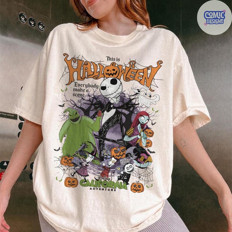 May include: Cream-colored t-shirt featuring a Halloween-themed graphic. The design includes Jack Skellington, Sally, Oogie Boogie, and other characters. The text on the shirt reads "This is Halloween" and "California Adventure".