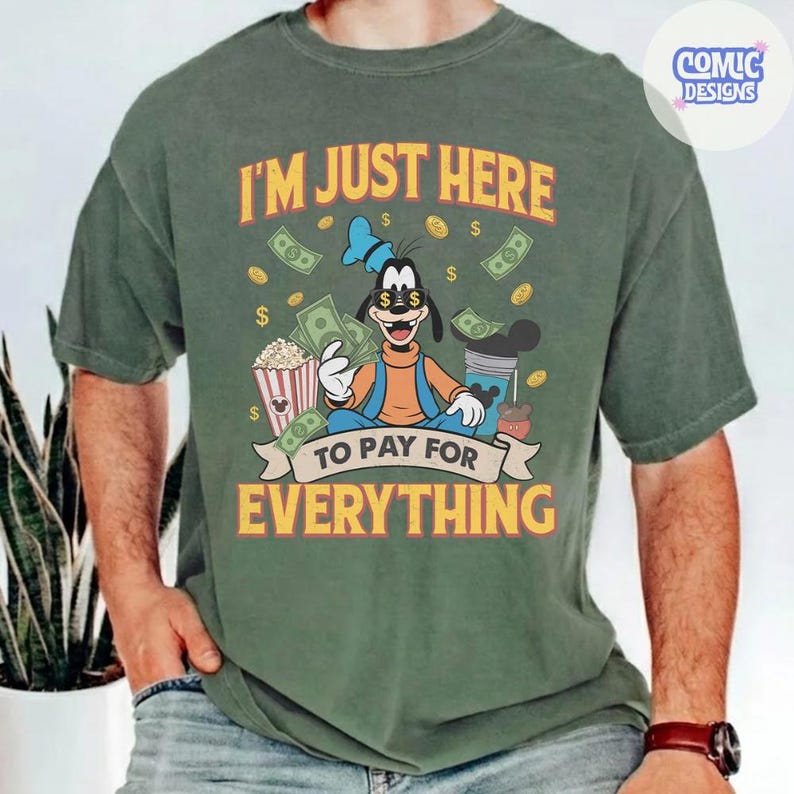 May include: Green t-shirt with a graphic of Goofy holding money, popcorn, and a drink. The text reads "I'M JUST HERE TO PAY FOR EVERYTHING" in yellow and orange. The shirt has short sleeves.