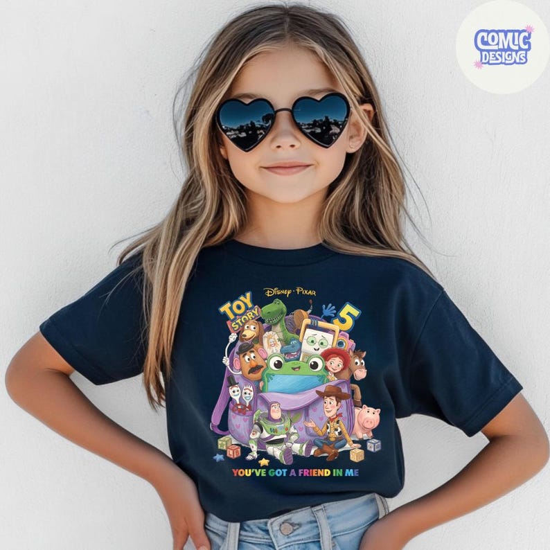 May include: A dark navy blue short-sleeve t-shirt featuring a colourful graphic print of Toy Story characters emerging from a purple backpack. The text reads Toy Story 5 and You've Got A Friend In Me. The shirt is worn with black heart-shaped sunglasses.
