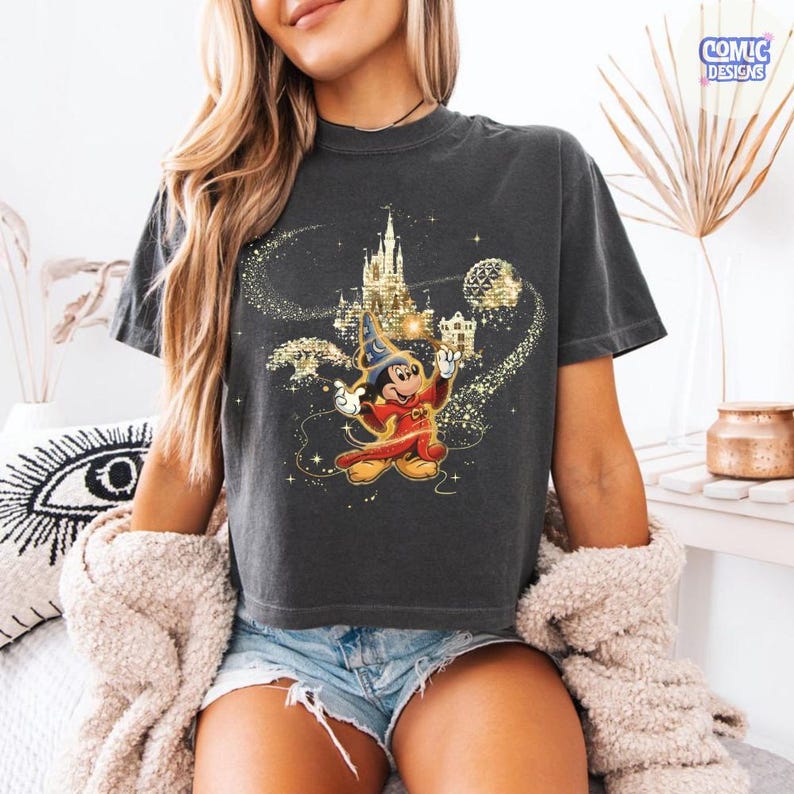 May include: Dark gray t-shirt featuring a graphic of Mickey Mouse in a sorcerer's outfit, with a castle and golden stars. The shirt has a crew neck and short sleeves. The Comic Designs logo is visible in the upper right corner.