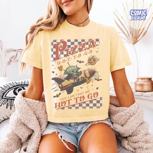 May include: A pale yellow short-sleeve t-shirt featuring a graphic of a green alien in a rocket ship. The design includes a checkered border, spiderwebs, bats, and the text Pizza H-O-T-T-O-G-O and You Can Take Me Hot To Go in a distressed font.
