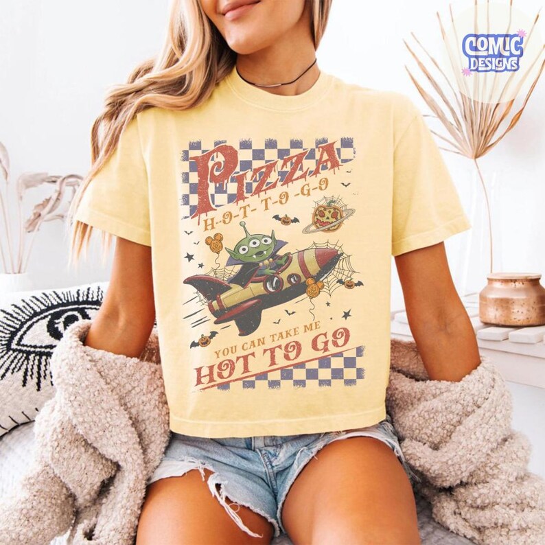 May include: A pale yellow short-sleeve t-shirt featuring a graphic of a green alien in a rocket ship. The design includes a checkered border, spiderwebs, bats, and the text Pizza H-O-T-T-O-G-O and You Can Take Me Hot To Go in a distressed font.