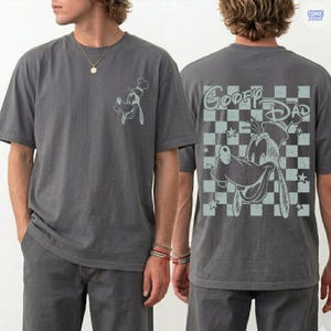May include: Gray t-shirt featuring a light blue graphic of Goofy wearing a chef's hat on the front. The back of the shirt displays Goofy with the text "Goofy Dad" on a checkered background. The shirt is short-sleeved.