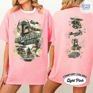 Puede incluir: Camiseta rosa claro con un dise&ntilde;o de la serie Star Wars, "The Mandalorian y Grogu." La parte delantera muestra el t&iacute;tulo y los personajes, mientras que la trasera presenta im&aacute;genes adicionales. La camiseta tiene el texto "This is the Way."