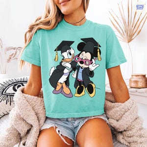 Puede incluir: Camiseta turquesa con un gr&aacute;fico de Daisy y Minnie Mouse con atuendo de graduaci&oacute;n. Los personajes de dibujos animados llevan birretes y togas. La camiseta es de color liso y tiene cuello redondo.