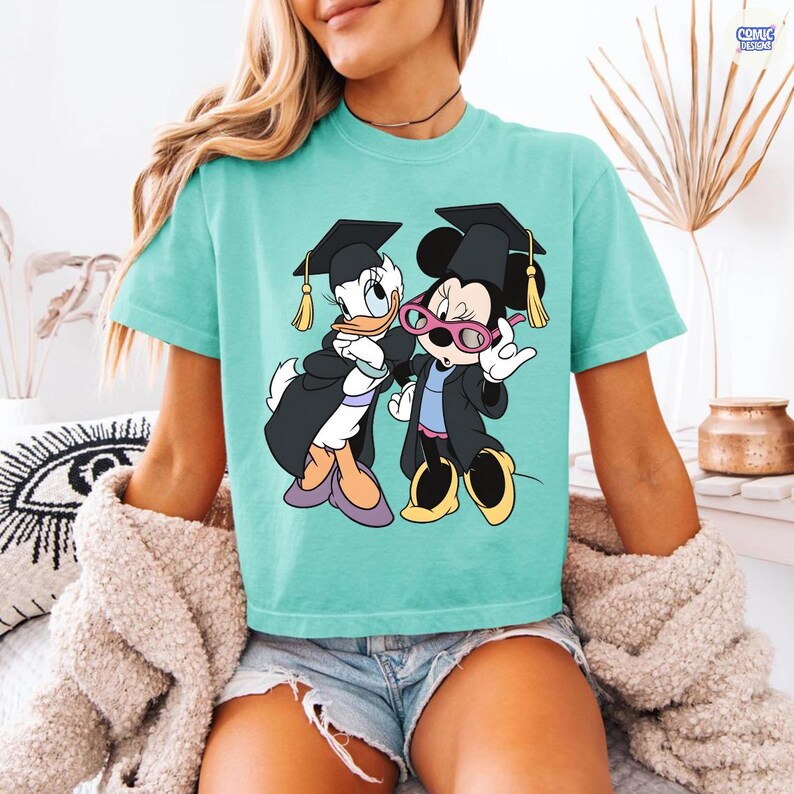 Puede incluir: Camiseta turquesa con un gr&aacute;fico de Daisy y Minnie Mouse con atuendo de graduaci&oacute;n. Los personajes de dibujos animados llevan birretes y togas. La camiseta es de color liso y tiene cuello redondo.