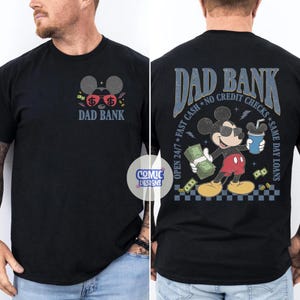 May include: Black t-shirt with a front graphic of Mickey Mouse ears with dollar signs and the words "DAD BANK". The back of the shirt features Mickey Mouse holding money and a drink, with text that reads "DAD BANK, Open 24/7, Fast Cash, No Credit Checks, Same Day Loans".