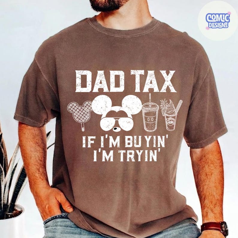 May include: Brown t-shirt with the text "DAD TAX" in white block letters. Below the text is a Mickey Mouse design with sunglasses, and food icons. The phrase "IF I'M BUYIN' I'M TRYIN'" is printed below the design.