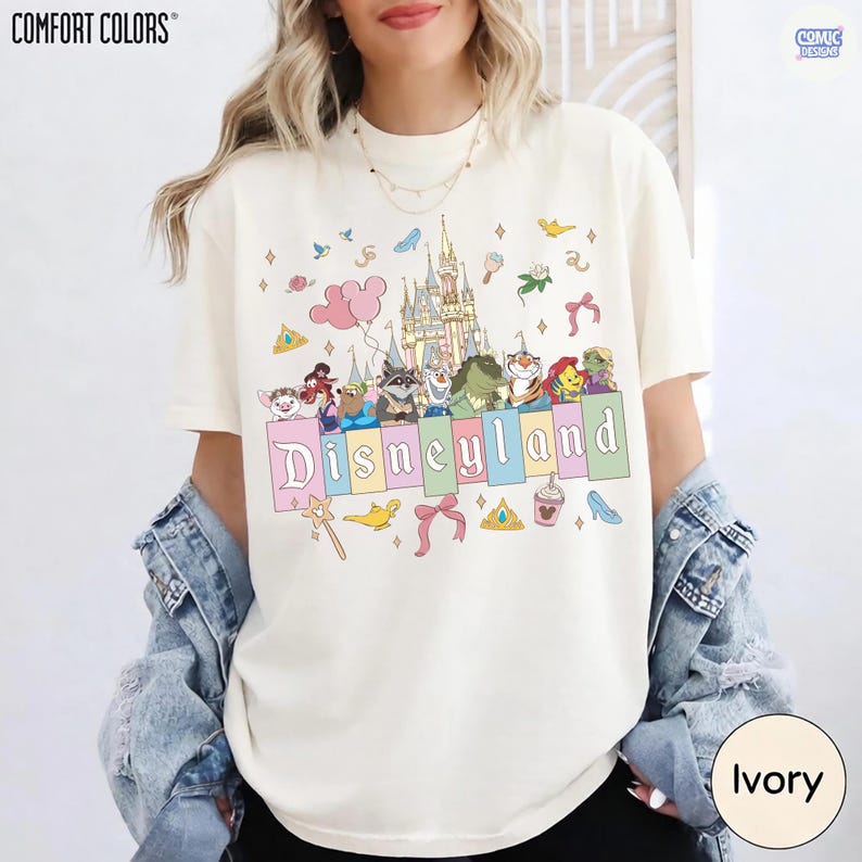 Pu&ograve; includere: T-shirt Comfort Colors color avorio con grafica Disneyland. Il design include un castello, personaggi dei cartoni animati e la scritta "Disneyland" in colori pastello. Grafiche aggiuntive includono una bacchetta, un fiocco e una scarpa con tacco.