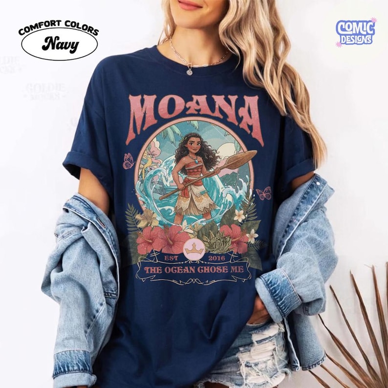May include: A navy blue t-shirt featuring a graphic print of the character Moana holding a wooden oar. The design includes tropical flowers, ocean waves, and the text MOANA, EST 2016, and THE OGEAN GHOSE ME. The shirt has a crew neckline and short sleeves.
