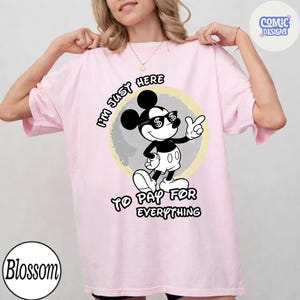 May include: Light pink t-shirt featuring a black and white Mickey Mouse graphic wearing sunglasses with dollar signs. The text reads "I'M JUST HERE TO PAY FOR EVERYTHING."