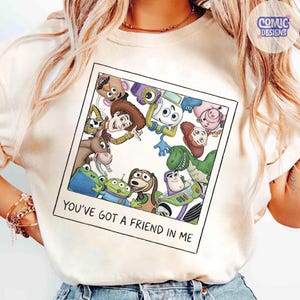 May include: A cream-colored t-shirt featuring a square graphic designed to look like a Polaroid photo. The print displays a group of animated characters from Toy Story, including Woody, Buzz Lightyear, and Rex, with the text YOU'VE GOT A FRIEND IN ME.