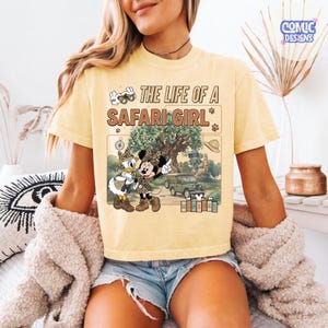 May include: Yellow t-shirt with the text "THE LIFE OF A SAFARI GIRL" and a graphic of Minnie and Daisy Duck in safari outfits. The graphic includes a jeep, tree, and safari hat. The shirt is a light yellow color.