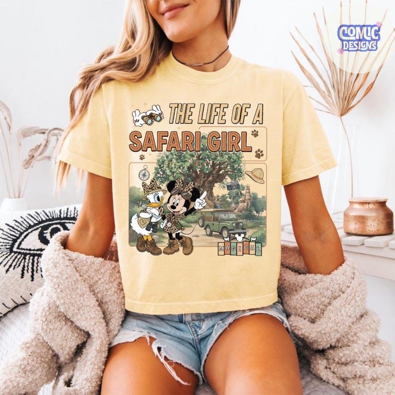 May include: Yellow t-shirt with the text "THE LIFE OF A SAFARI GIRL" and a graphic of Minnie and Daisy Duck in safari outfits. The graphic includes a jeep, tree, and safari hat. The shirt is a light yellow color.