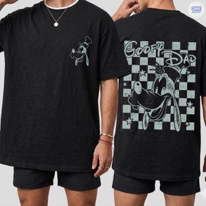 May include: Black t-shirt featuring a cartoon image of Goofy wearing a chef's hat on the front and back. The back of the shirt has the words "Goofy Dad" in a checkered pattern with stars. The shirt is made of a soft material.