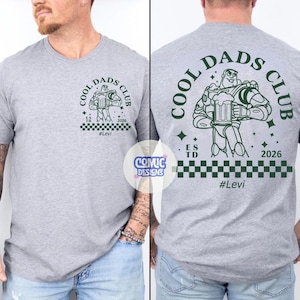 May include: A light gray t-shirt with a green graphic of Buzz Lightyear holding a beer mug, with the text "COOL DADS CLUB" and "#Levi". The back of the shirt also features the same design. The shirt has a checkered pattern at the bottom.
