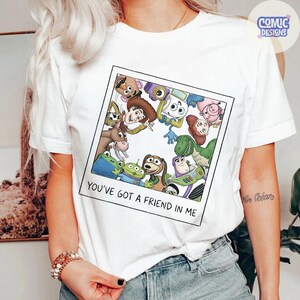 May include: A white short-sleeve t-shirt featuring a square graphic print of various cartoon characters from the Toy Story franchise arranged in a circle. Below the characters, the text reads, "YOU'VE GOT A FRIEND IN ME" in black capital letters.