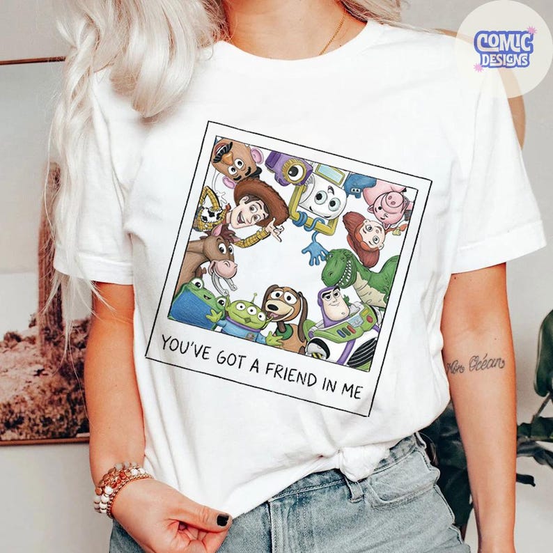 May include: A white short-sleeve t-shirt featuring a square graphic print of various cartoon characters from the Toy Story franchise arranged in a circle. Below the characters, the text reads, "YOU'VE GOT A FRIEND IN ME" in black capital letters.