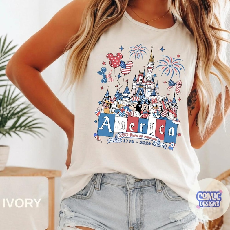 May include: An ivory sleeveless tank top featuring a graphic print of a castle, fireworks, and cartoon characters in red, white, and blue. The text reads America, 250 Years of Freedom, and 1776-2026, with Mickey Mouse-shaped balloons and stars.
