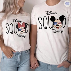 May include: Matching white t-shirts with the text "Disney Squad". One shirt features a Minnie Mouse design with a red bow and the word "Mommy". The other shirt has a Mickey Mouse design and the word "Daddy".