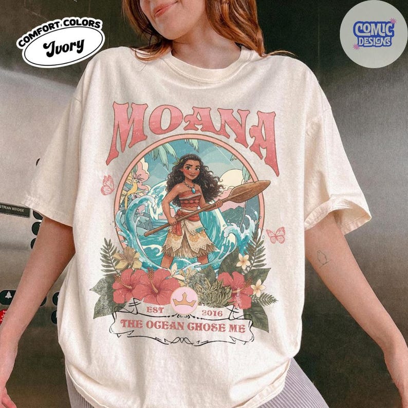 May include: An ivory t-shirt featuring a graphic of the character Moana holding a wooden oar. The design includes a circular frame, blue ocean waves, red hibiscus flowers, and the text MOANA, EST 2016, and THE OGEAN GHOSE ME.