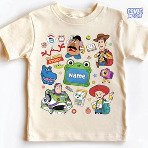 May include: A cream-colored short-sleeve t-shirt featuring a colorful graphic print of various cartoon characters including a space ranger, a cowboy, a cowgirl, a potato figure, and a spork. The text 1ST GRADE STORY and NAME are clearly visible.