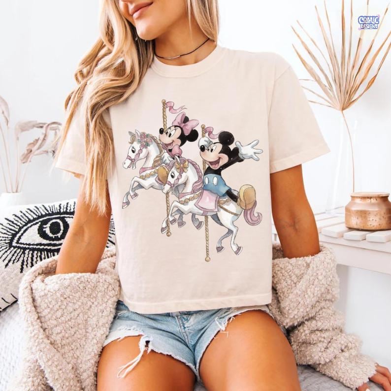 May include: Cream-colored t-shirt featuring a carousel design with Mickey Mouse and Minnie Mouse riding white horses. The graphic includes pink and blue accents. The shirt is worn by a person.