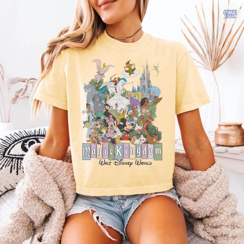 Puede incluir: Camiseta amarilla clara con un gr&aacute;fico colorido de personajes de Disney, incluyendo Mickey Mouse y Cenicienta. El texto "Magic Kingdom Walt Disney World" est&aacute; impreso debajo. La camiseta tiene mangas cortas.