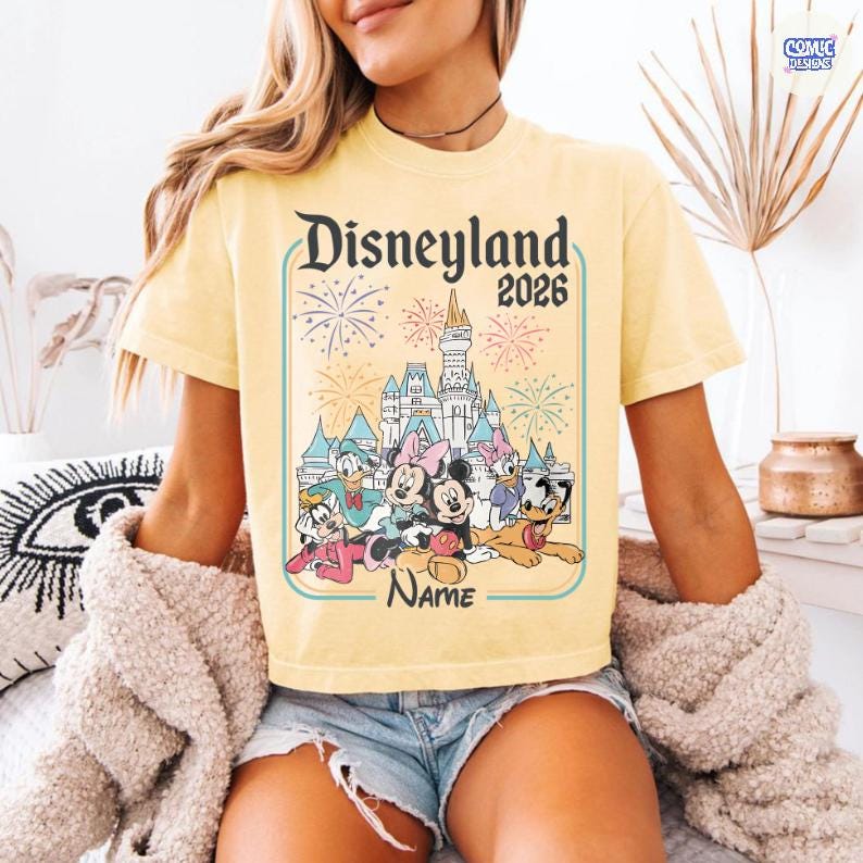 Op de afbeelding: Lichtgeel T-shirt met een Disneyland 2026-afbeelding. Het ontwerp bevat een kasteel, vuurwerk en tekenfilmfiguren zoals Mickey en Minnie Mouse. Het T-shirt heeft een lichtgele kleur.