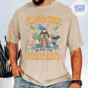 May include: Beige t-shirt with a graphic of a cartoon character holding money, popcorn, and a drink. The text on the shirt reads "I'M JUST HERE TO PAY FOR EVERYTHING". The design includes dollar bills and coins.