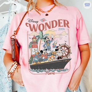 Puede incluir: Camiseta rosa con un dise&ntilde;o de personajes de Disney en un crucero, el texto dice "Disney Wonder 2026". El dise&ntilde;o incluye a Mickey Mouse, Pato Donald, Goofy y Daisy. Tambi&eacute;n se ve un faro y palmeras.