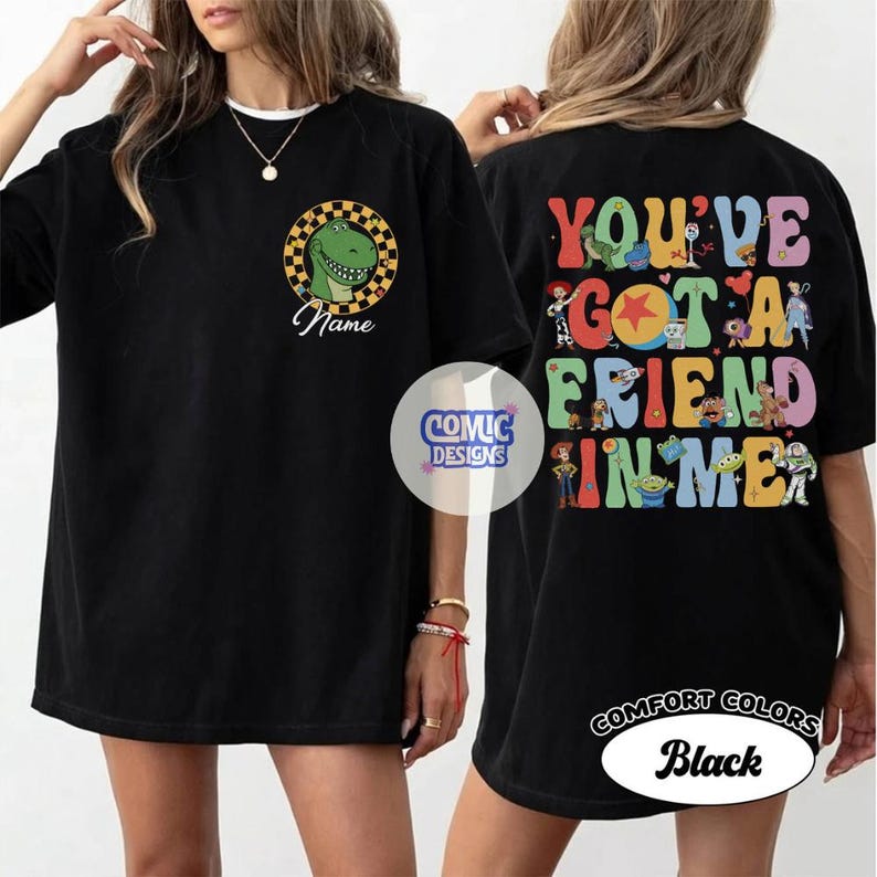 Op de afbeelding: Een zwart t-shirt met een afbeelding op de voorkant van een groene dinosaurus in een geel en zwart geblokte cirkel met het woord Name eronder. De achterkant toont de kleurrijke tekst YOU'VE GOT A FRIEND IN ME met diverse stripfiguren.