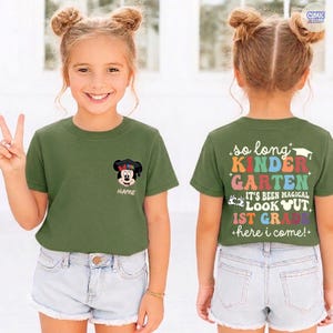 May include: Olive green t-shirt with a Mickey Mouse graphic wearing a graduation cap. The front of the shirt has the word "NAME" below the graphic. The back of the shirt says "so long KINDERGARTEN" and "1st GRADE".
