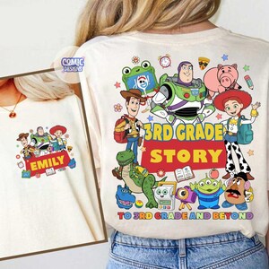 May include: A cream-colored t-shirt featuring a colorful graphic of Toy Story characters including Woody, Buzz Lightyear, Jessie, Rex, and Mr. Potato Head. The text reads 3RD GRADE STORY TO 3RD GRADE AND BEYOND with the name EMILY printed.