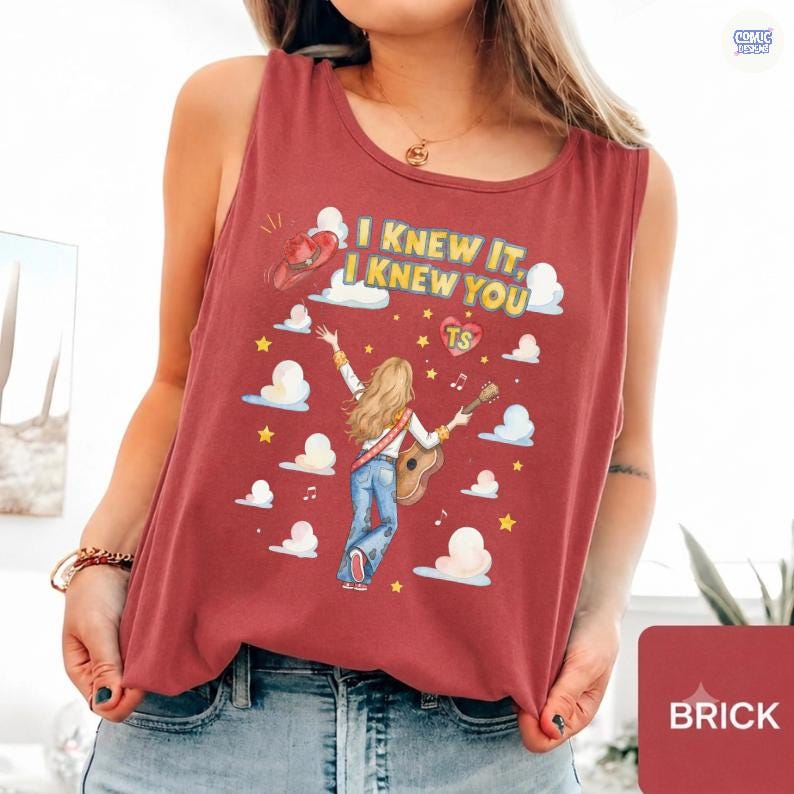 May include: A brick red sleeveless tank top featuring a graphic print of a person with long blonde hair playing an acoustic guitar. The text reads I KNEW IT, I KNEW YOU, with a heart containing TS, a cowboy hat, clouds, stars, and musical notes.