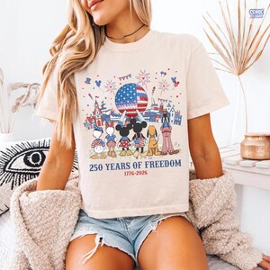 May include: A cream-colored t-shirt featuring a graphic of cartoon characters standing before a geodesic dome and fireworks. The text reads 250 YEARS OF FREEDOM 1776-2026. The design includes red, white, and blue patriotic motifs and star patterns.