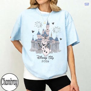 May include: Light blue t-shirt featuring a graphic of a castle, fireworks, and Olaf the snowman. The text "Disney Trip 2026" is printed below the graphic. The shirt has short sleeves and a crew neck.