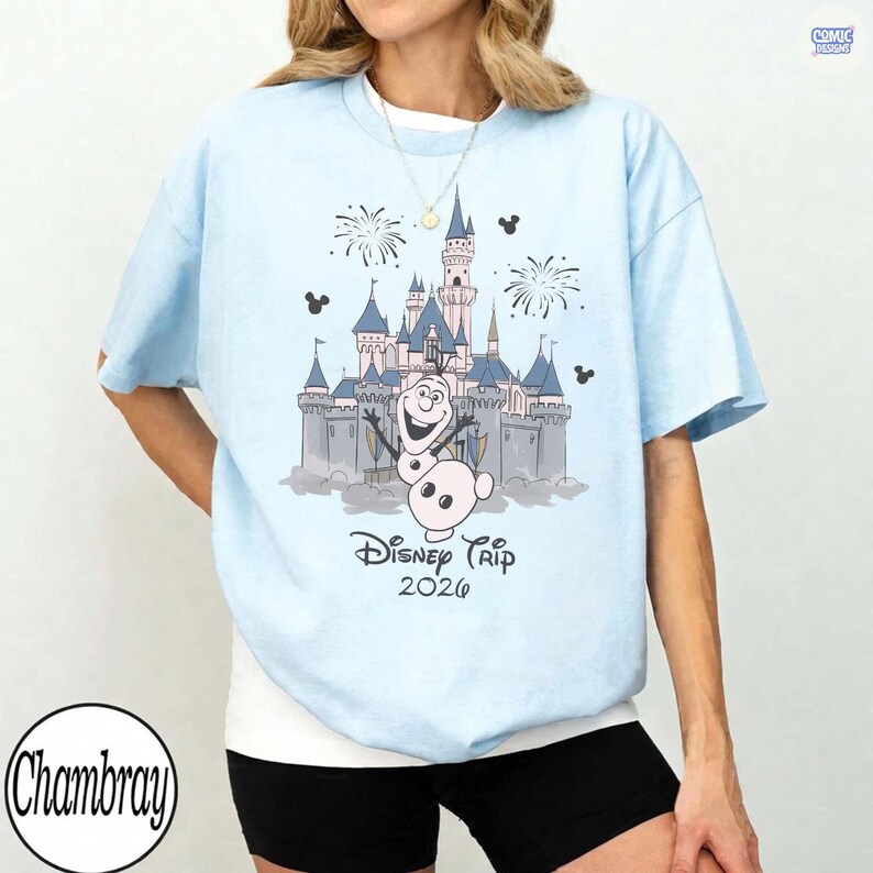 May include: Light blue t-shirt featuring a graphic of a castle, fireworks, and Olaf the snowman. The text "Disney Trip 2026" is printed below the graphic. The shirt has short sleeves and a crew neck.