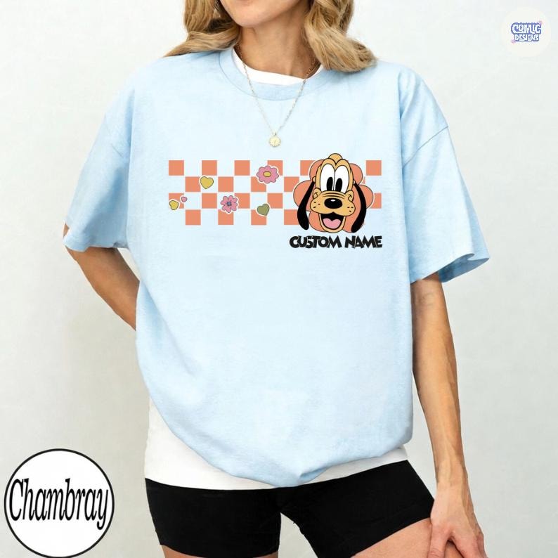 May include: Light blue t-shirt featuring a cartoon dog graphic with a custom name option. The design includes a checkered pattern with flowers and hearts. The shirt is a casual wear item.