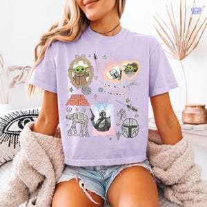 Puede incluir: Camiseta lila claro con ilustraciones de Star Wars. El dise&ntilde;o incluye a Grogu, el Mandaloriano y la frase "THIS IS THE WAY". La camiseta es de corte relajado, adecuada para el uso diario.